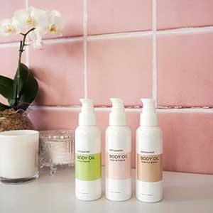 Three white bottles of body oil in front of a pink tile wall