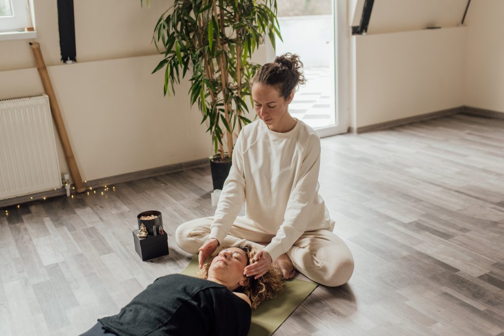 A woman lies comfortably on the floor, surrounded by an array of crystals, while a natural therapist stands beside her, offering Reiki energy healing. The woman, with closed eyes, wears a peaceful expression on her face. The therapist, dressed in earth-toned attire, holds their hands above the woman's body, directing healing energy towards her.